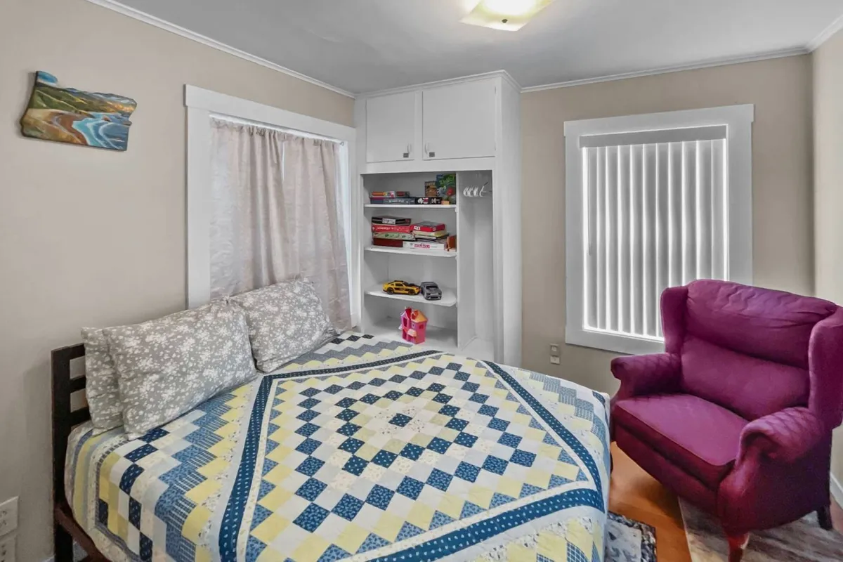 A cozy bedroom scene featuring a bed with a quilt, a pink armchair in the corner, and a shelf holding various items. The space is warm and welcoming, clearly designed for relaxing with friends, reading, or gathering for game nights.