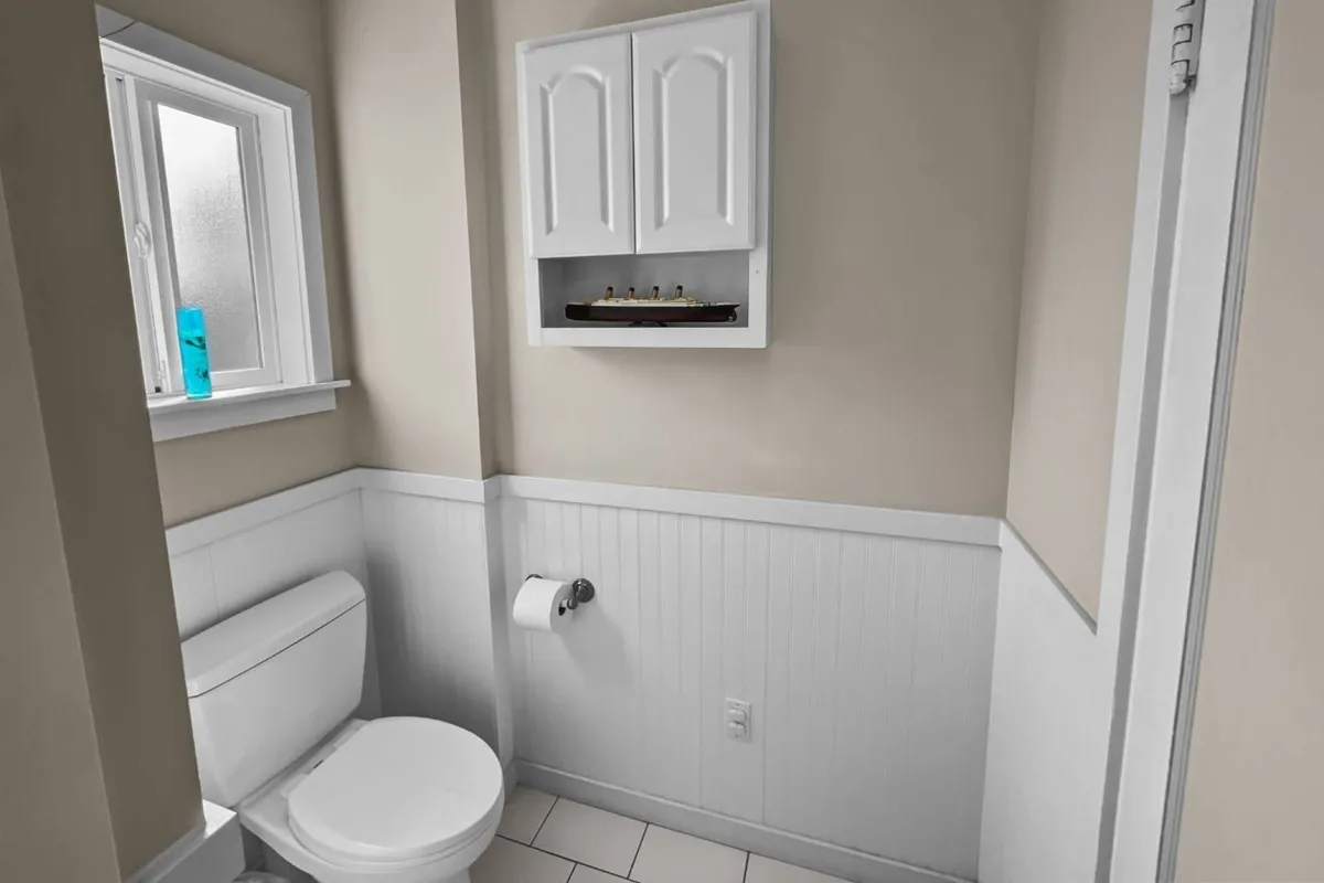 A compact and practical bathroom corner is shown, featuring a window that filters in natural light, a decorative shelf with a model ship, and thoughtfully arranged towels. The scene feels cozy and subtly nautical, hinting at the cottage's vintage charm.