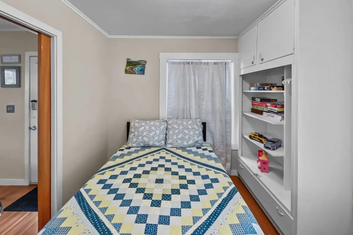 A flexible sleeping space featuring a bed with a cozy quilt, a nearby bookshelf, and a window with a curtain that adds softness to the space. The inviting mood makes it suitable for relaxing, reading, or accommodating extra guests, capturing the flexible loft theme.