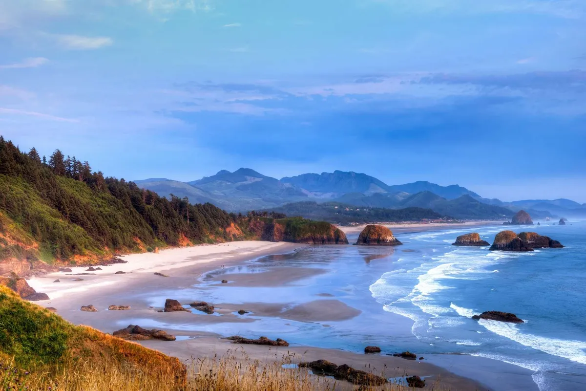 A scenic exterior shot of a sandy beach with ocean waves, rocky formations, distant mountains, and a tree line. The wide view evokes the feeling of nearby seaside adventure, aligning perfectly with the section's theme of launching into coastal explorations from home.