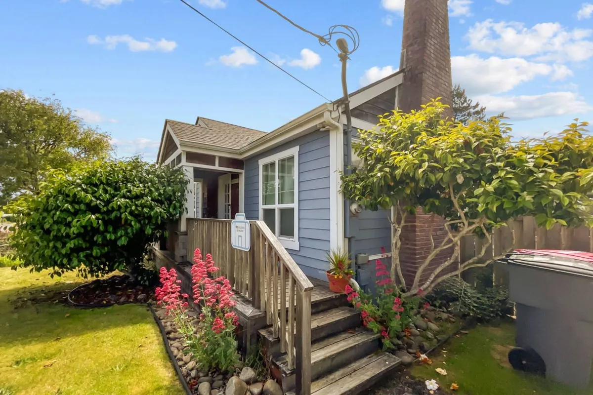 The image showcases the exterior of a charming single-family cottage with inviting blue siding, a welcoming front porch, colorful flower beds in full bloom, and a classic brick chimney. The cozy porch is perfect for relaxing, and the lush garden hints at the joys of outdoor gatherings or quiet moments in nature.