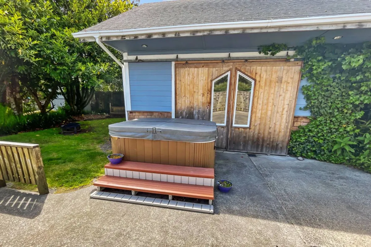 This exterior image focuses on a hot tub set against a backdrop of natural greenery, with a wooden door in the background. The area conveys a private oasis vibe, perfect for relaxing under the open sky. The lush surroundings suggest a tranquil space for both solo and group enjoyment.