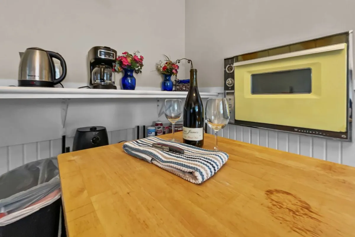 A warmly lit kitchen and dining space featuring a sturdy wooden table set with a bottle of wine and glasses, a retro oven, coffee maker, kettle, and a bright bouquet of colorful flowers. The scene radiates sociability and culinary potential, while details like the wine and flowers highlight the space’s readiness for both intimate dinners and festive gatherings.