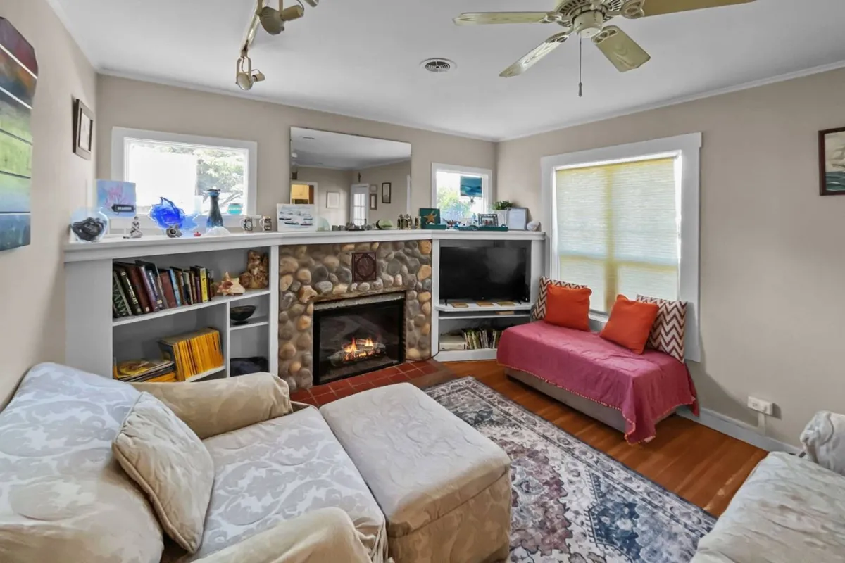 A bright, inviting living room with a plush sofa facing a streaming TV and a cozy fireplace. Bookshelves and a window bring natural light and charm, while coastal décor and comfortable seating create a relaxing, communal atmosphere ideal for family gatherings and movie nights.