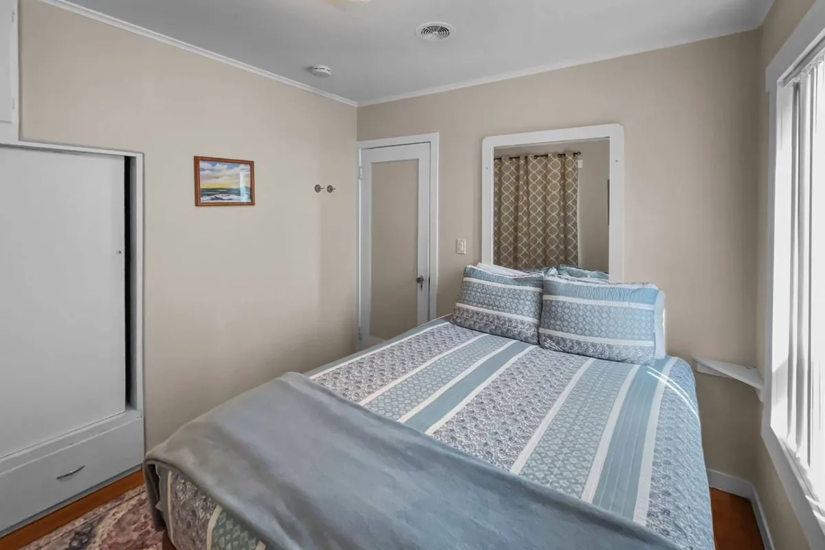 A bright bedroom with a bed dressed in patterned bedding, framed artwork above, and a window letting in ample natural light. The room exudes an inviting and serene vibe, ideal for quiet moments or starting the day refreshed.