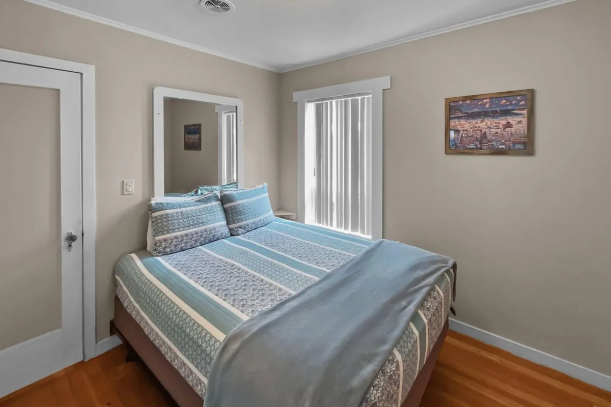 A cozy bedroom featuring a bed adorned with decorative pillows, a mirror that reflects natural light into the space, and a painting on the wall. The scene has an inviting and tranquil atmosphere, ideal for rest and relaxation.